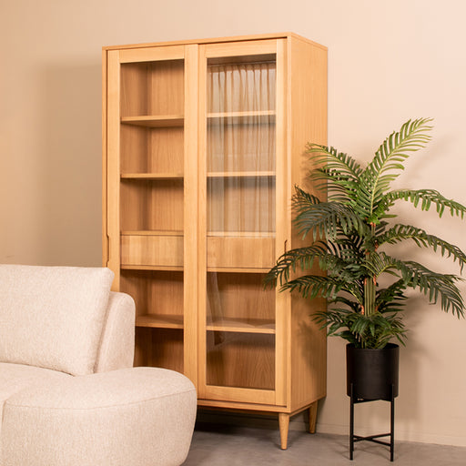 LABEL51 Jule vitrinekast van naturel eikenhout met glazen deuren, 100x49x180 cm, in een modern interieur.