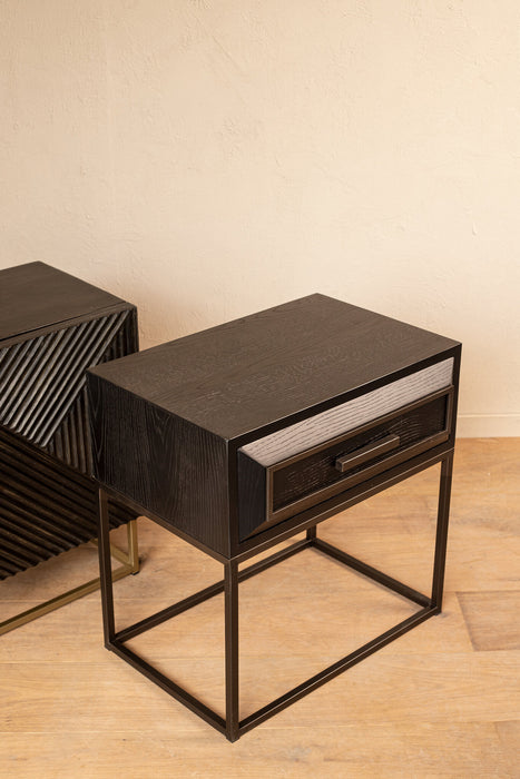 Zwarte PTMD Lixly opbergkast van ijzer met één lade en een slank metalen onderstel op een houten vloer.